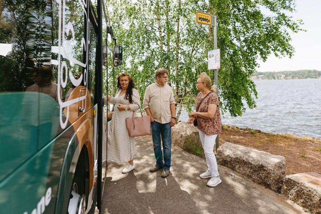 joukko kesäisiä ihmisiä nousee bussin kyytiin meren äärellä Meripuistossa